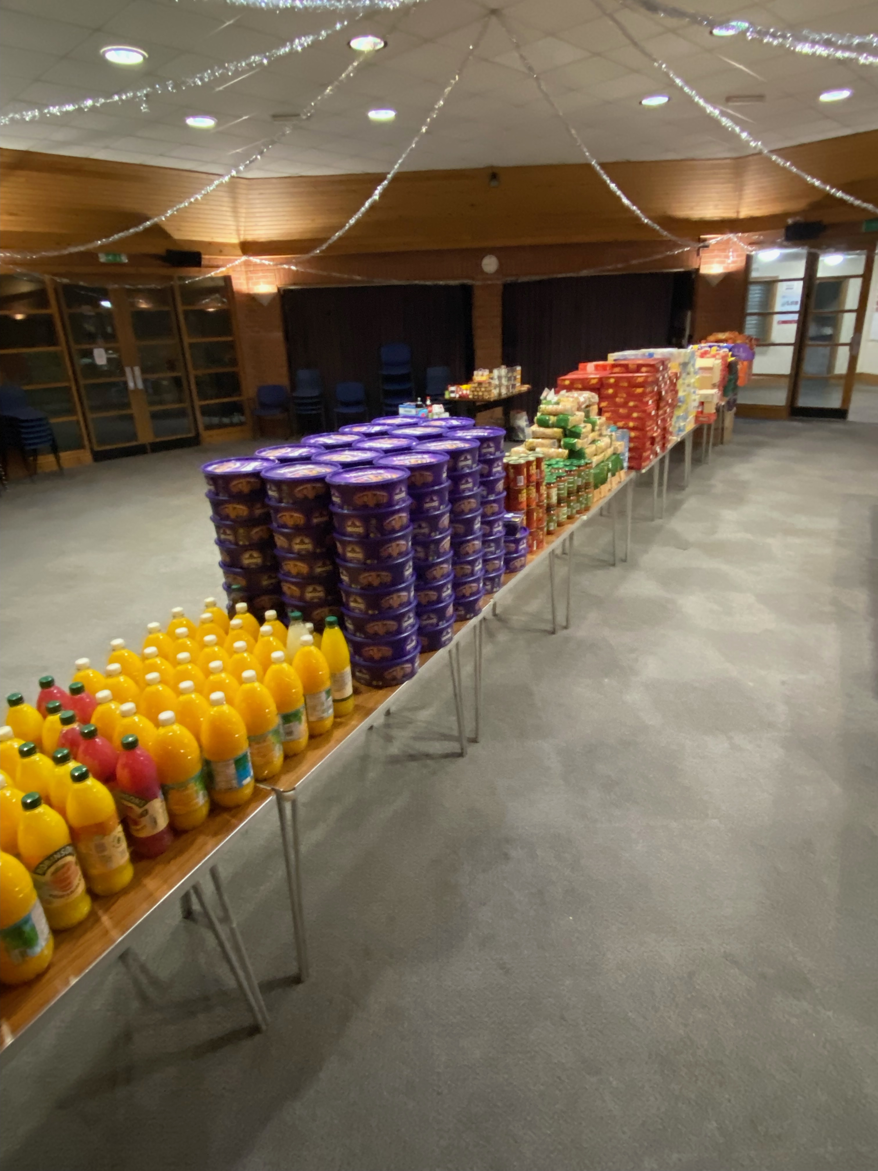 Tables with stackes of squash bottles, chocolate boxes, crips packets and biscuits, waiting to be packed into gift boxes